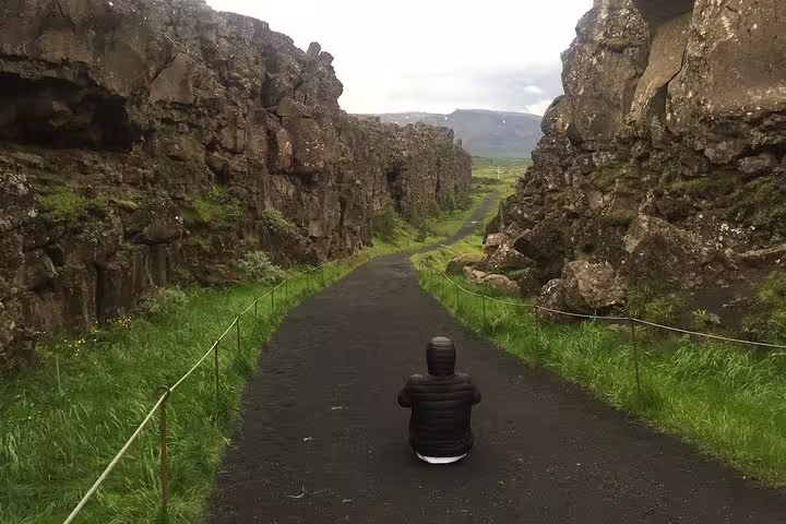 Person sitting on black lava rock path between towering cliffs at Þingvellir National Park, Iceland