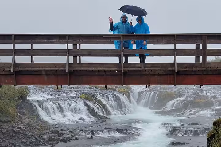 Two tourists in blue rain ponchos with umbrella on bridge over waterfall during Golden Circle tour