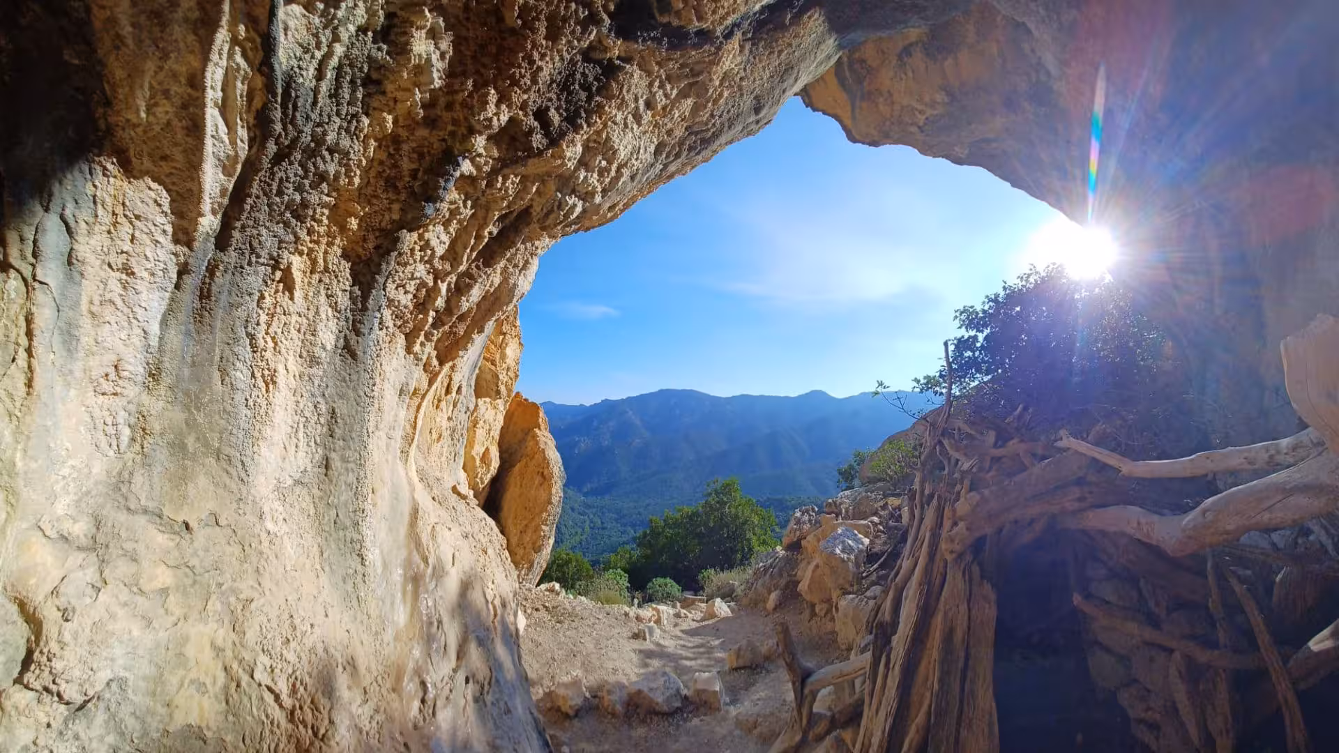 Sunlight streaming through a rocky cave entrance with panoramic views of Gorropu canyon's lush landscape.