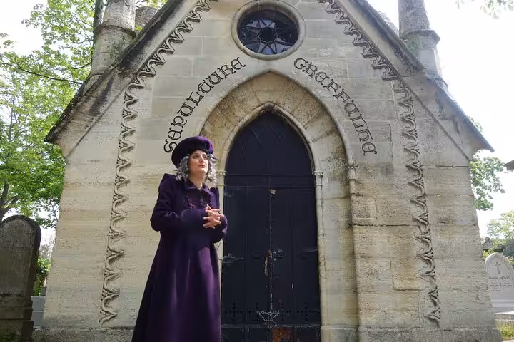 Woman in purple Victorian coat and hat stands before gothic stone mausoleum at Pere Lachaise Cemetery