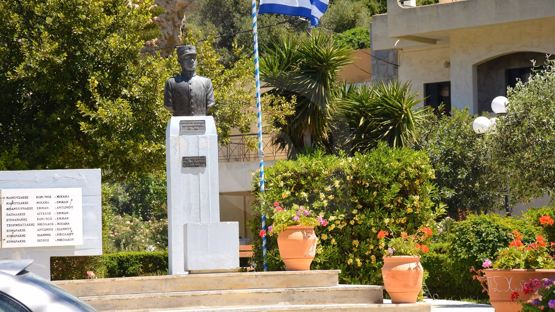 Greek village memorial statue with flag and flower pots, scenic stop on all-inclusive 2-day tour with overnight stay