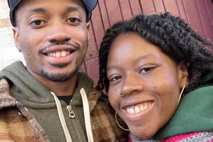 Two smiling people posing for a selfie outdoors near a red wooden building in Greenwich Village