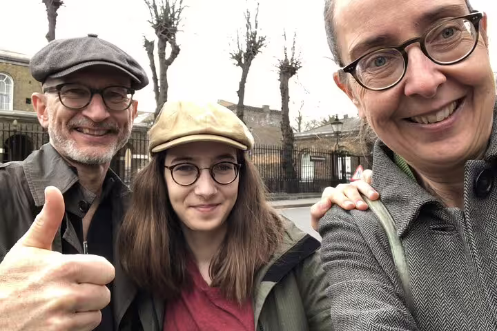 Three smiling visitors on a self-guided walking tour in Greenwich streets, two wearing flat caps and glasses
