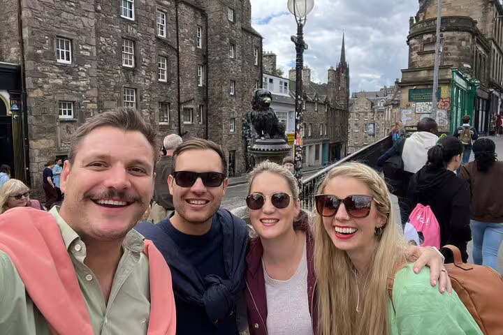 Four smiling tourists posing near Greyfriars Bobby statue on Edinburgh's historic Candlemaker Row