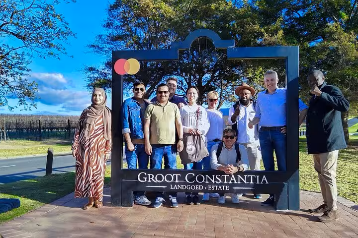 Group of tourists posing at the iconic Groot Constantia estate entrance surrounded by picturesque vineyards.