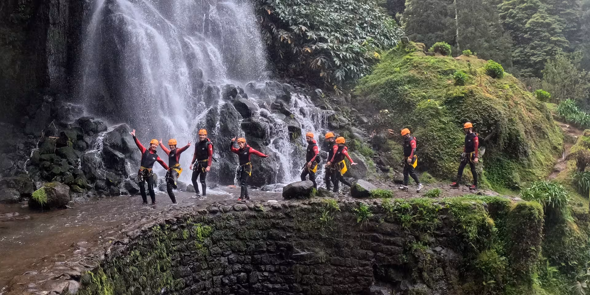 Group canyoning tour in wetsuits and helmets walking beside a powerful waterfall on an outdoor adventure route
