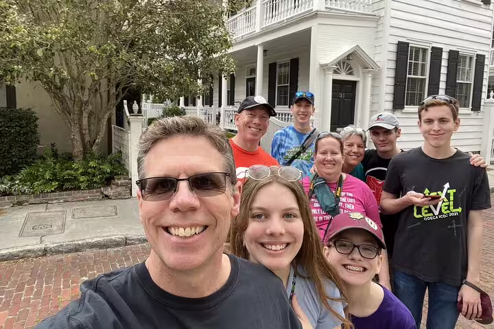 Group of tourists smiling for selfie on brick street in front of historic white Charleston home