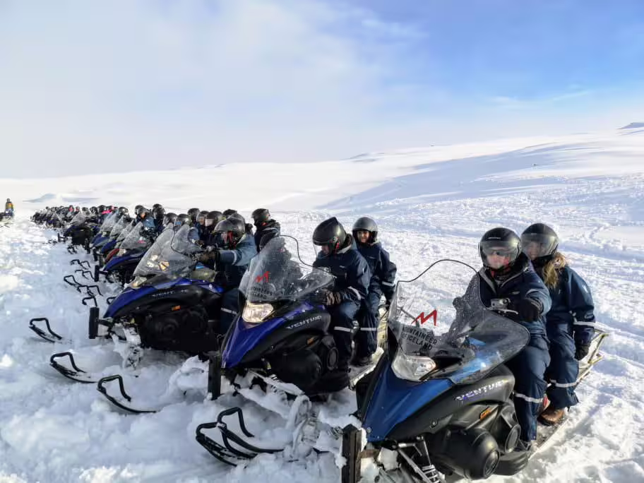 A group of snowmobilers in helmets and suits lined up on blue snowmobiles across a vast Iceland glacier
