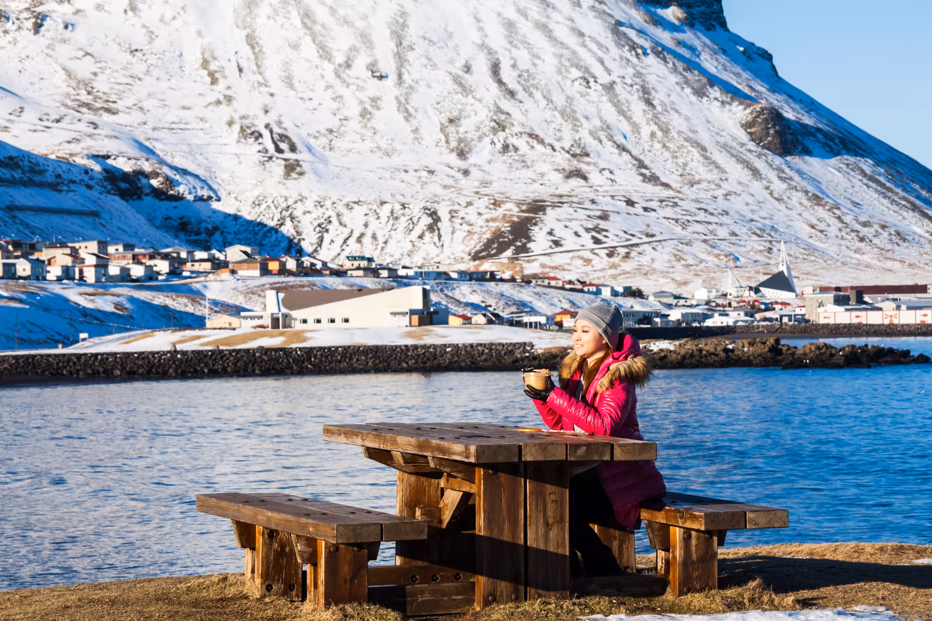 Woman in pink jacket enjoying hot drink at picnic table by Grundarfjordur harbor with snowy Snaefellsnes mountains
