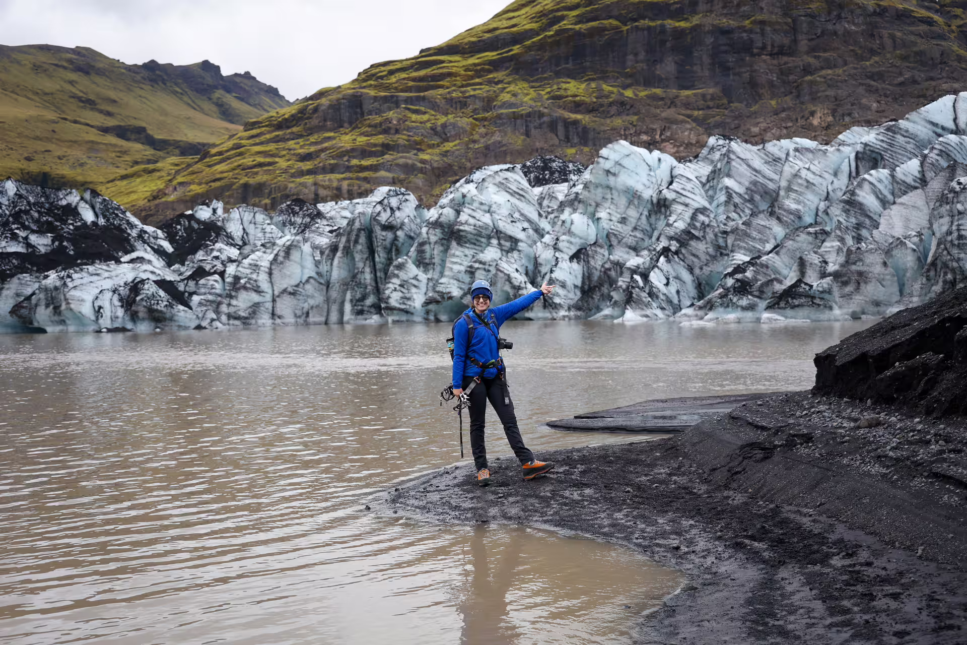 A smiling guide in blue jacket points toward the stunning Solheimajokull glacier ice wall in Iceland.