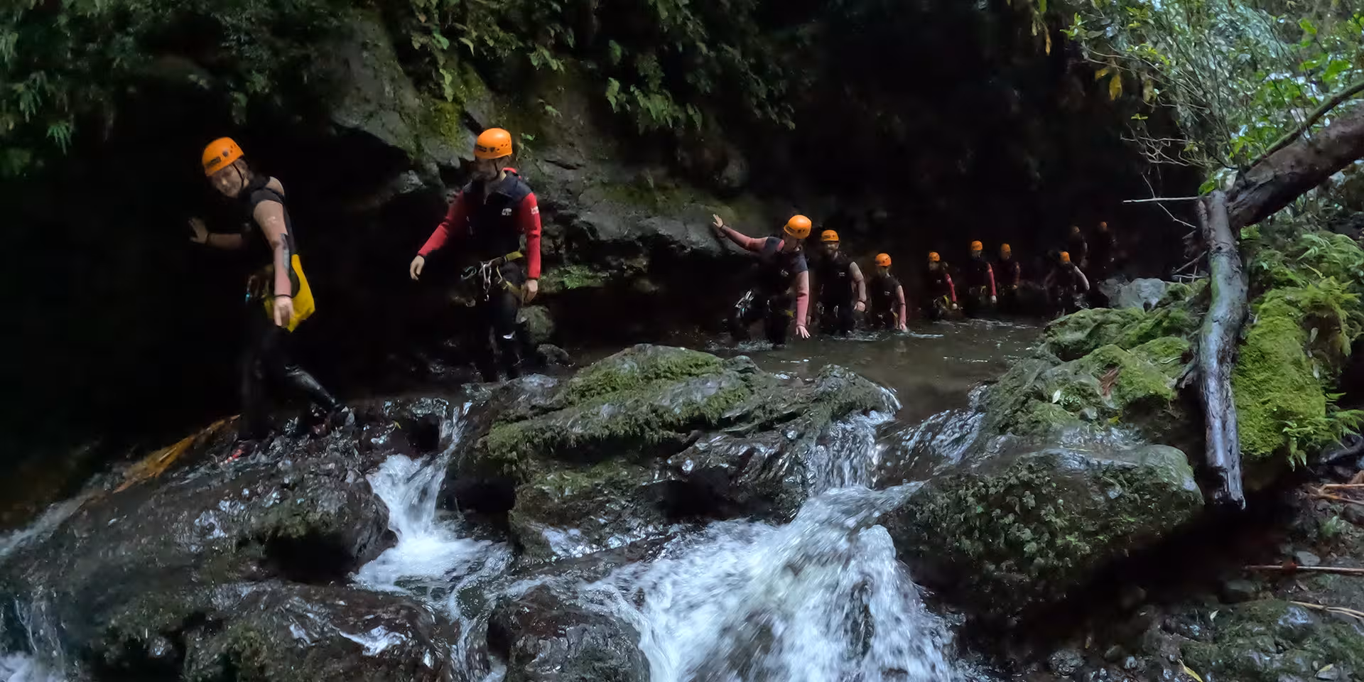 Guided canyoning group trekking through a mossy river gorge in helmets and wetsuits, adventure tour