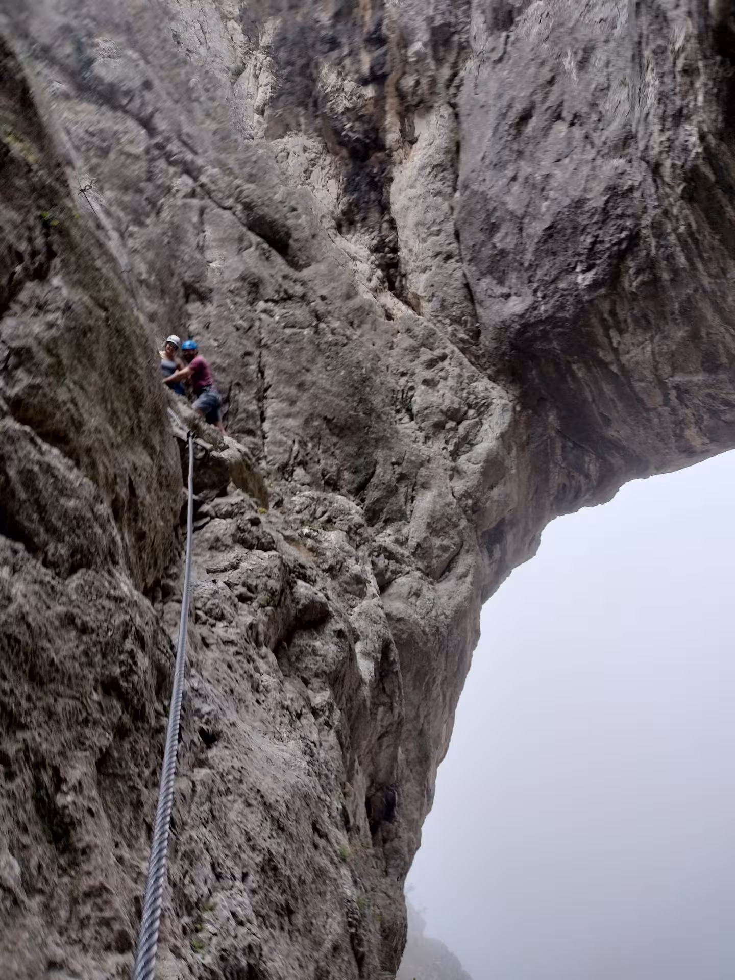 Two hikers with helmets climbing rocky cliff face in Turda Canyon with steel safety cable