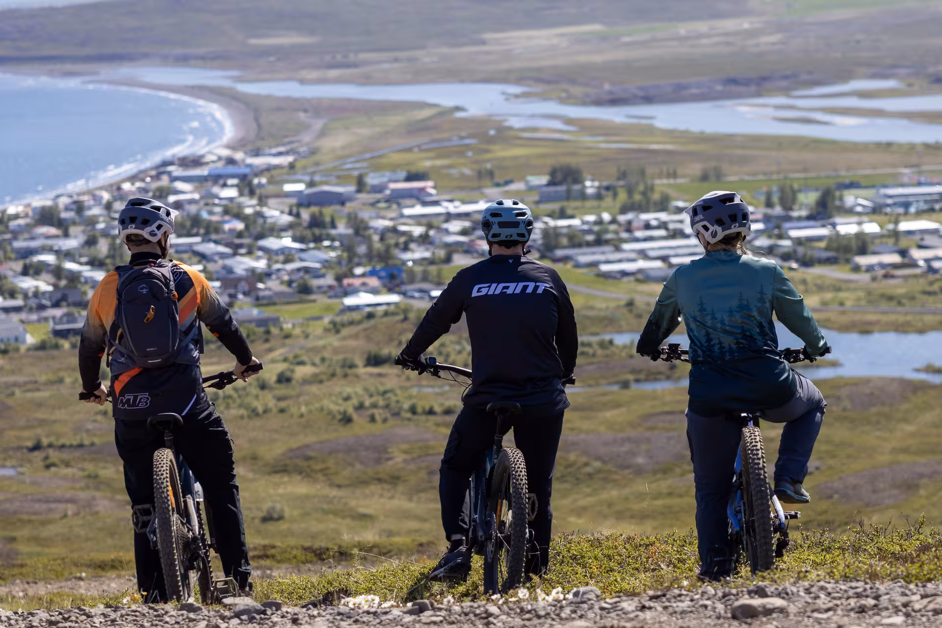 Three mountain bikers in helmets pause on scenic hillside overlooking coastal town during guided e-MTB tour