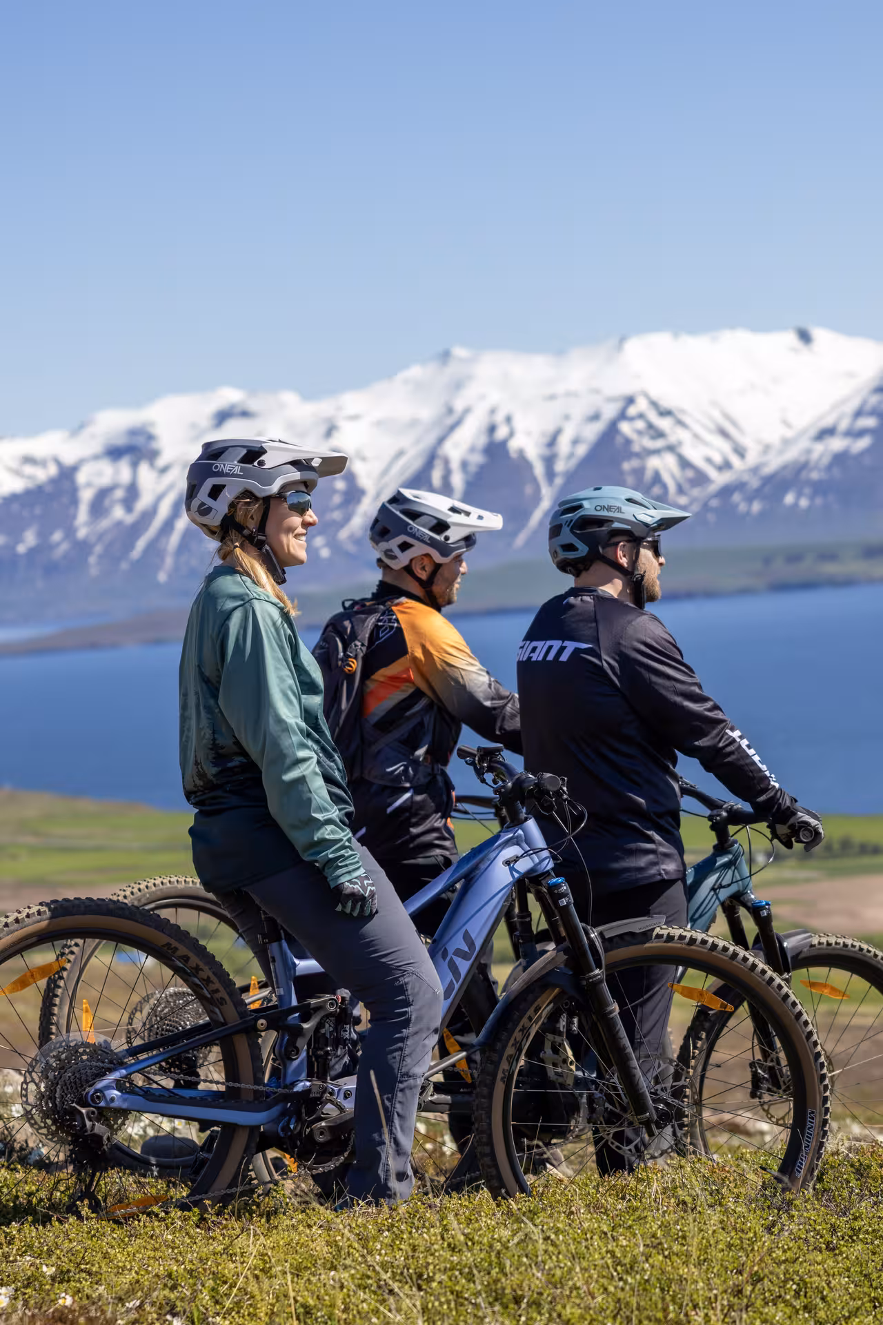 Three mountain bikers with helmets pause on scenic hilltop overlooking lake and snow-capped mountains on e-MTB tour