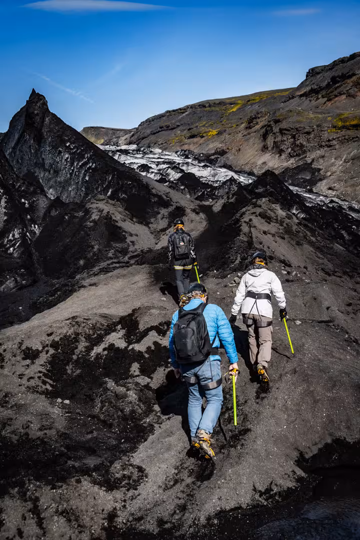 Three hikers with trekking poles walking across black volcanic glacier terrain at Solheimajokull Iceland
