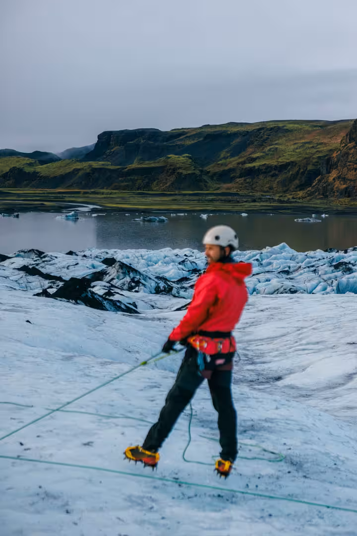 Guide in red jacket and crampons rappelling on Solheimajokull glacier with glacial lagoon behind