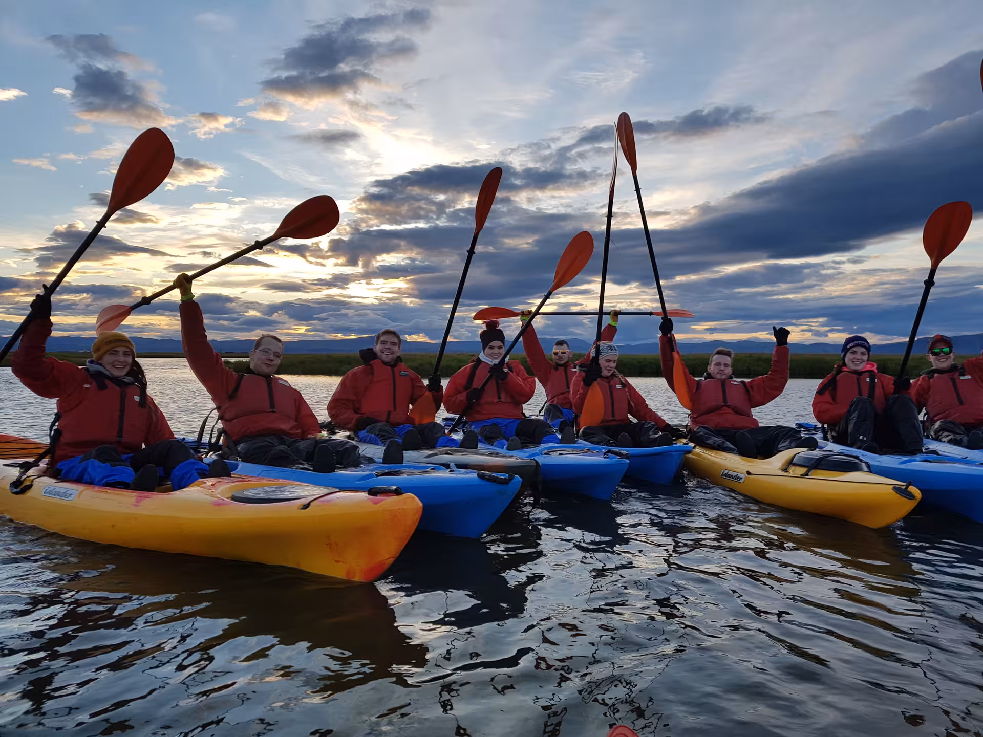 Group of kayakers raising paddles in colorful kayaks on calm Icelandic waters at sunset