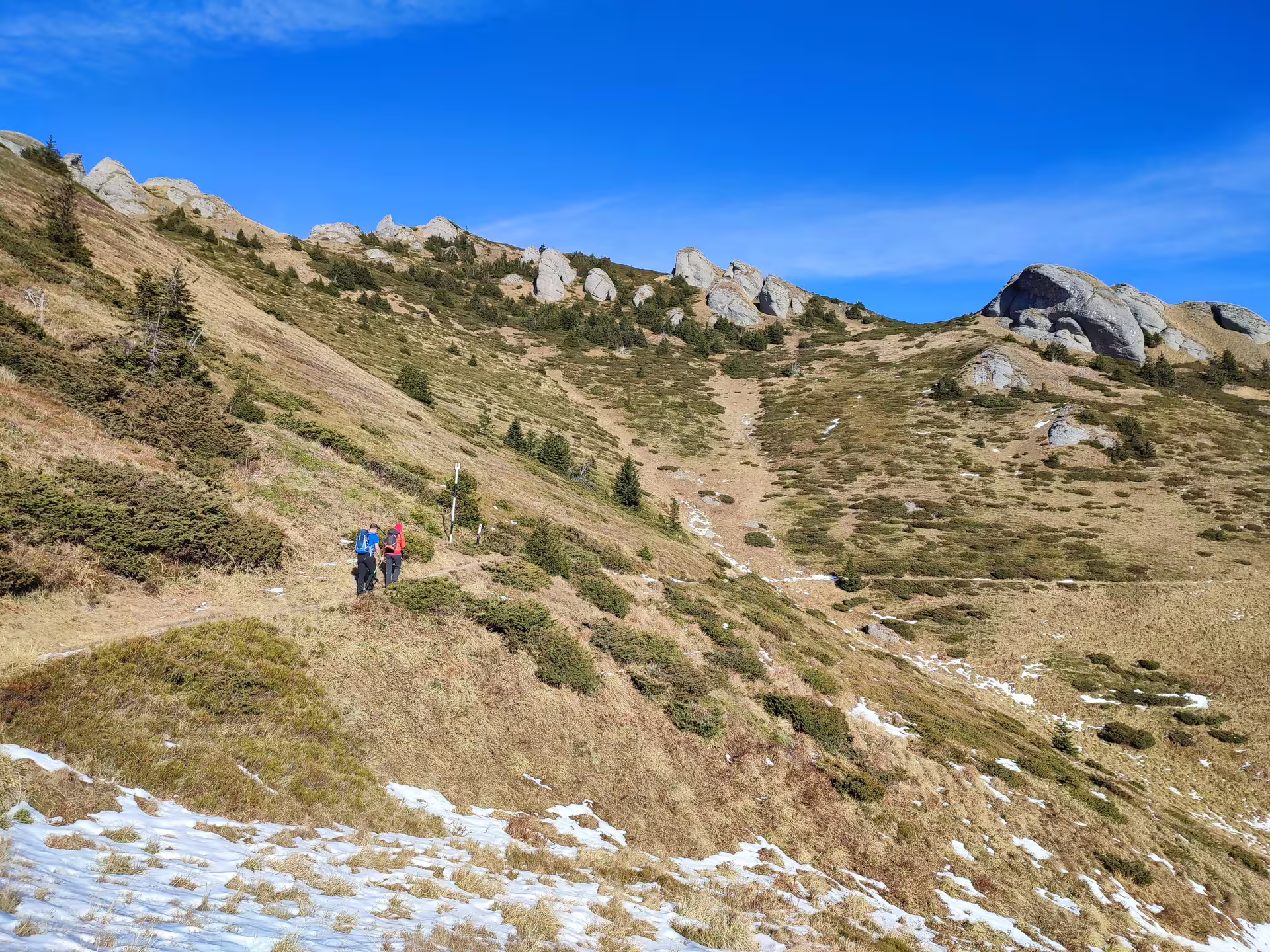 Hikers on trail toward Ciucas Peak rocky summit, Romania, with snow patches and alpine meadows