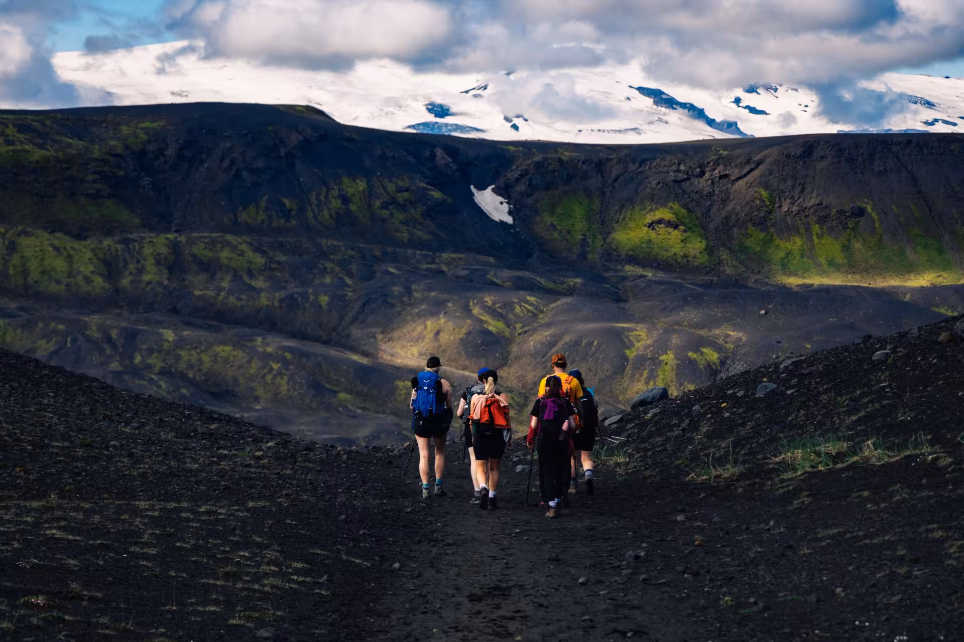 Group of hikers trekking on black volcanic trail with snow-capped mountains in Iceland