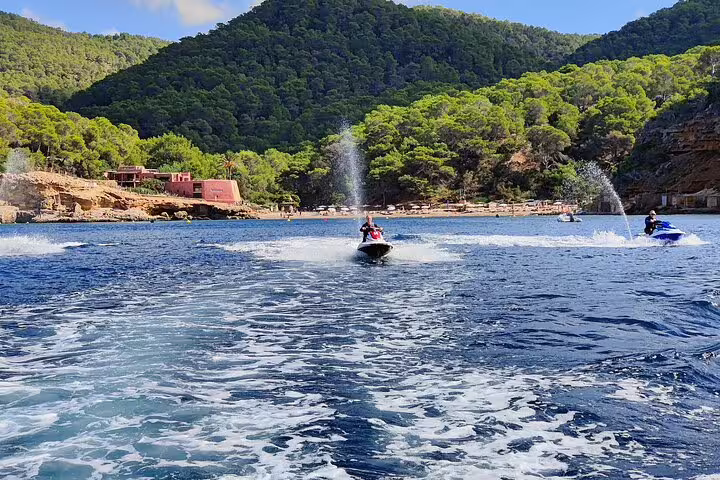 Guided jet ski tour to Cala Salada Ibiza, riders speeding across turquoise bay with pine-covered cliffs