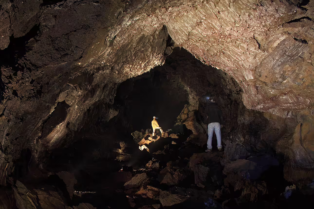 Two explorers with headlamps standing inside a massive lava cave tunnel with rocky floor