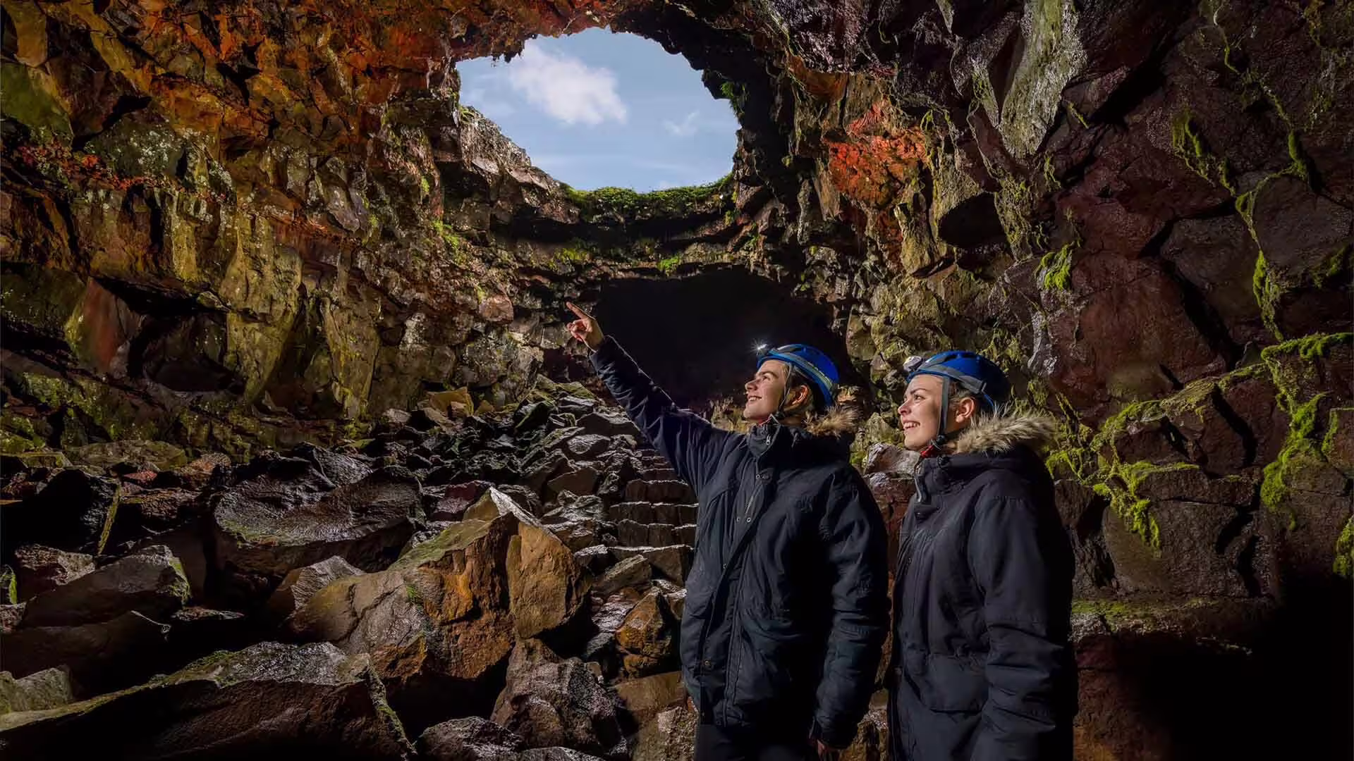 Two explorers wearing blue helmets inside an Icelandic lava tunnel pointing at a skylight opening above