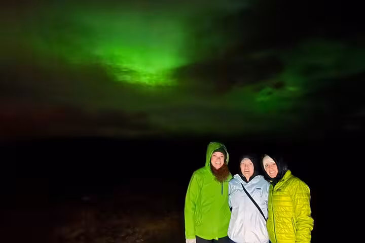 Three women smiling under vibrant green northern lights aurora borealis in Reykjavik night sky