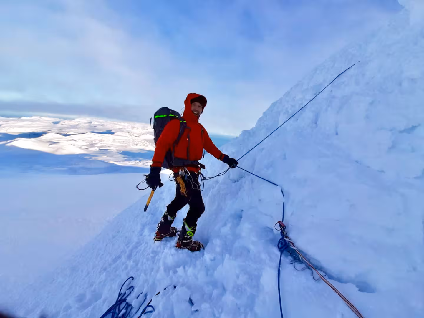 Smiling mountaineer in red jacket climbing snowy ridge at Snaefellsjokull glacier with rope and crampons