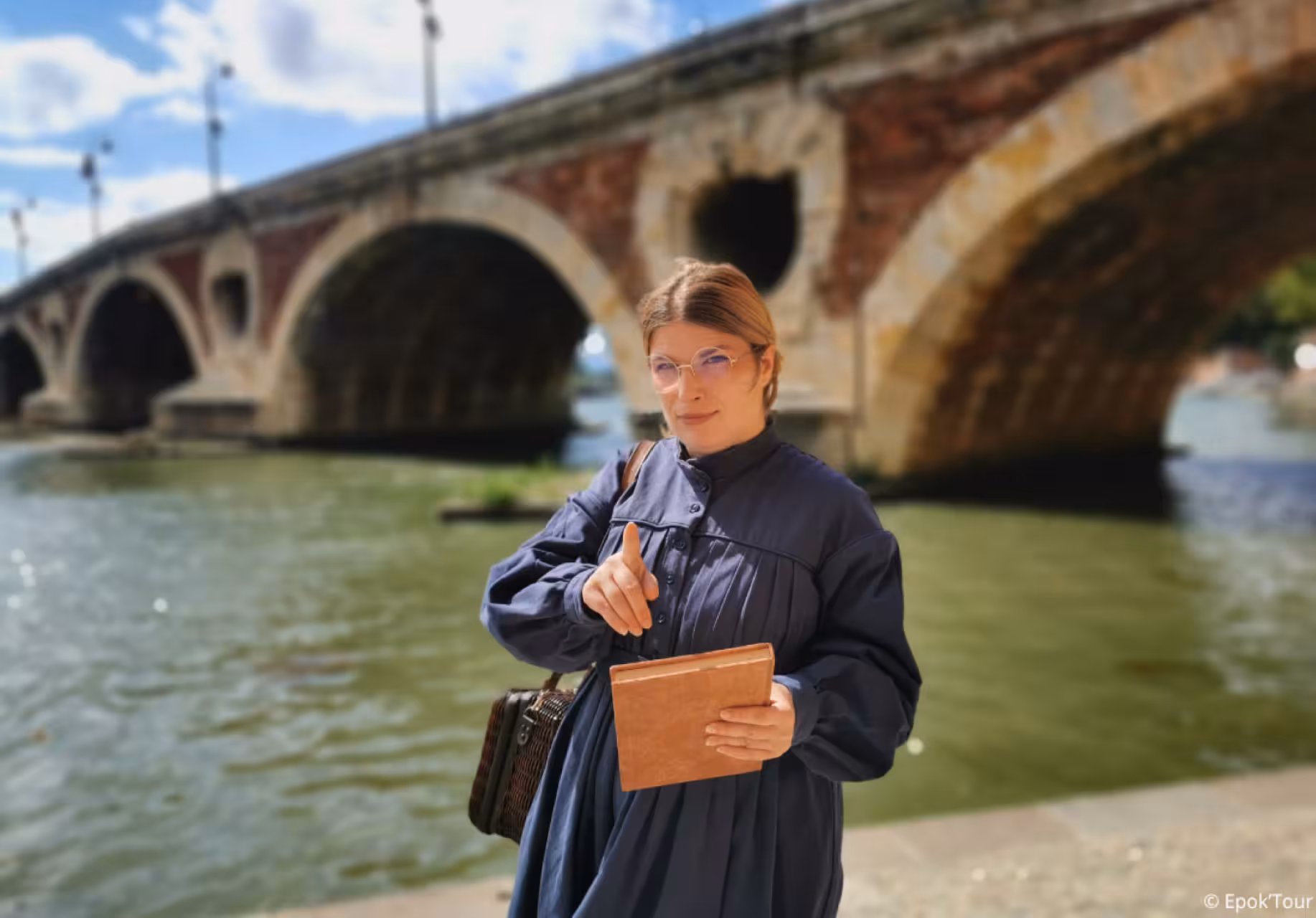 Female guide in navy dress holding a book by the Garonne River with Pont Neuf bridge in Toulouse