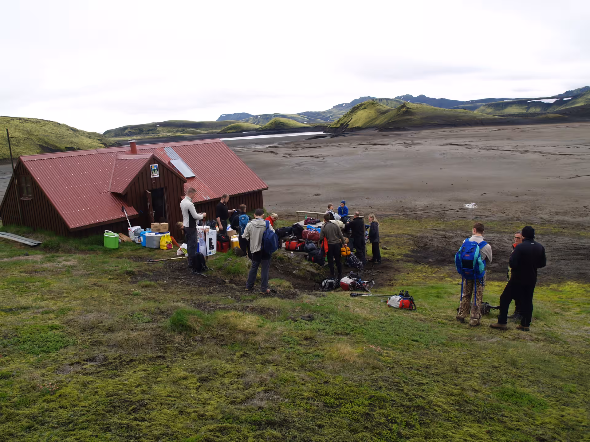 Hikers gathering with gear outside a red mountain hut near Eldgja Canyon, Iceland's highlands in background