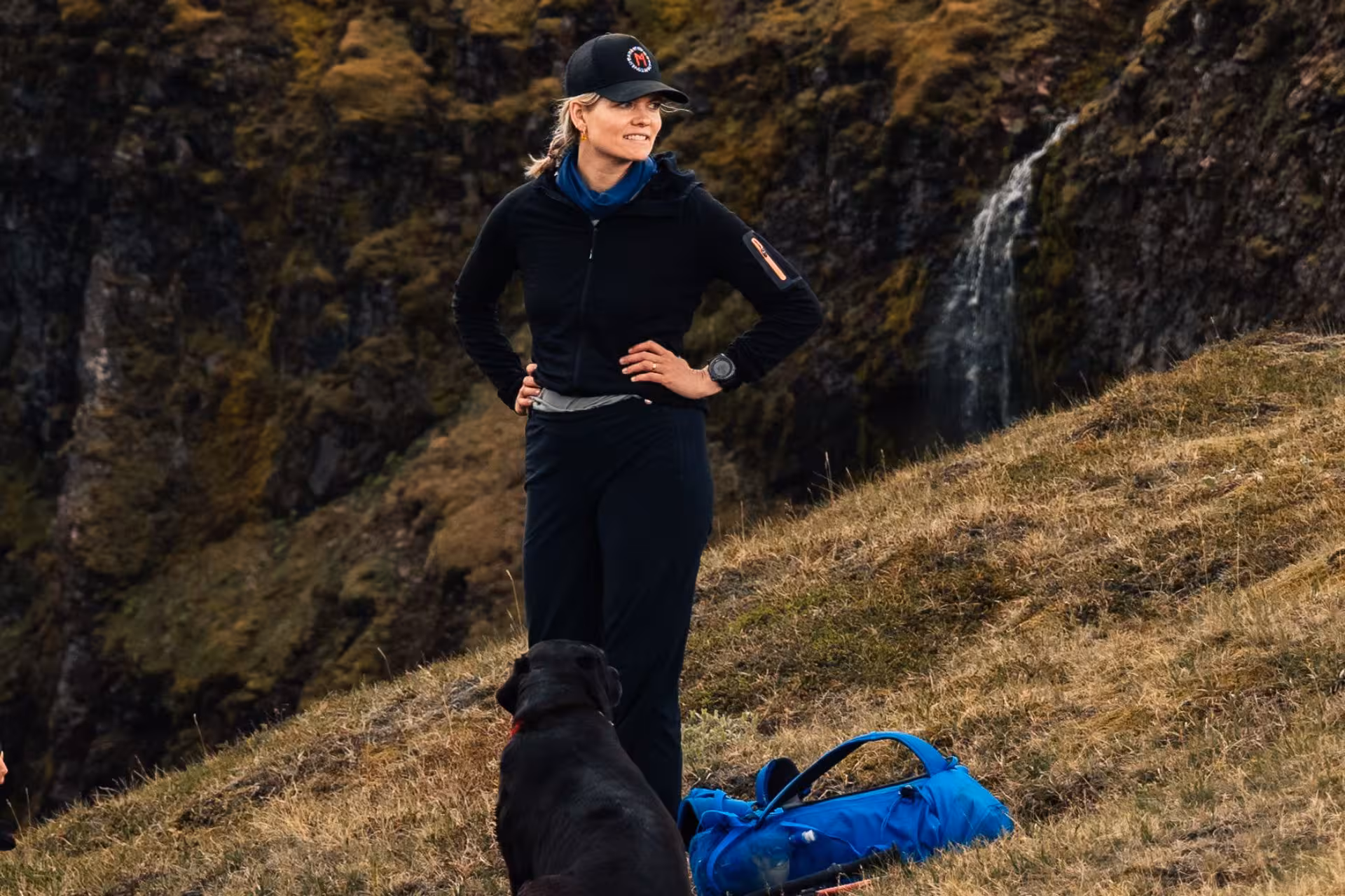 Woman hiker in black gear standing on grassy Icelandic hillside with black dog and waterfall backdrop