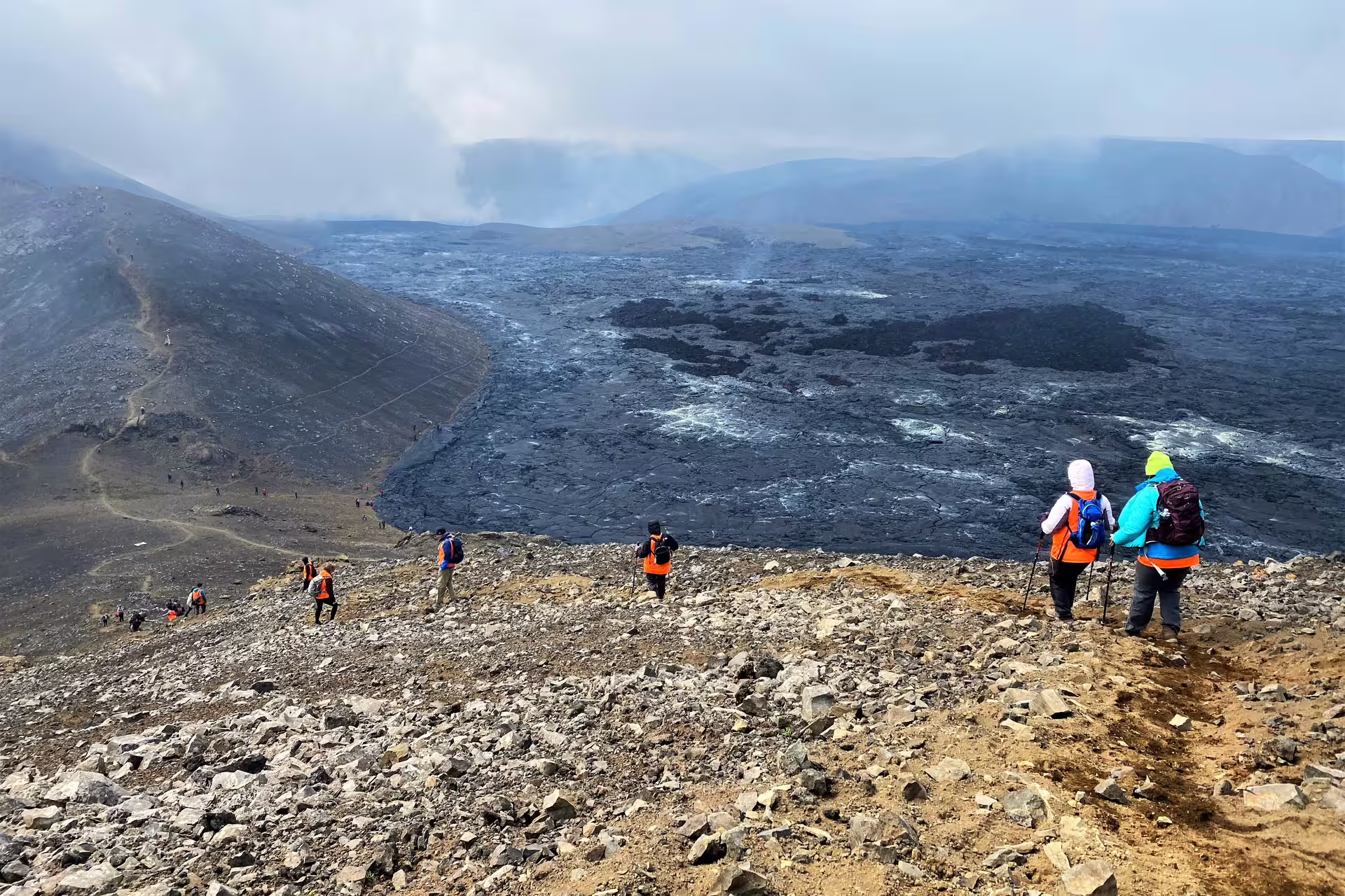Hikers in orange vests descending toward a vast black lava field on the Reykjanes Peninsula, Iceland