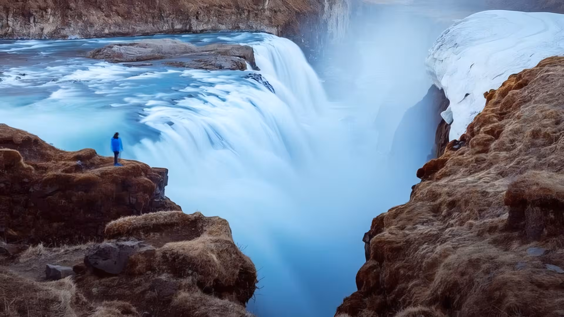 Person in blue jacket standing on rocky cliff overlooking Gullfoss waterfall in Iceland in winter