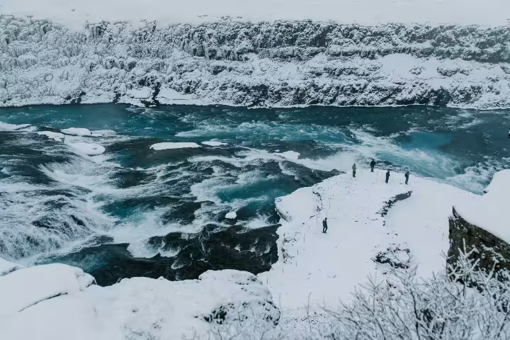 Tourists standing on snow-covered rocky ledge beside turquoise rapids at Gullfoss waterfall Iceland in winter