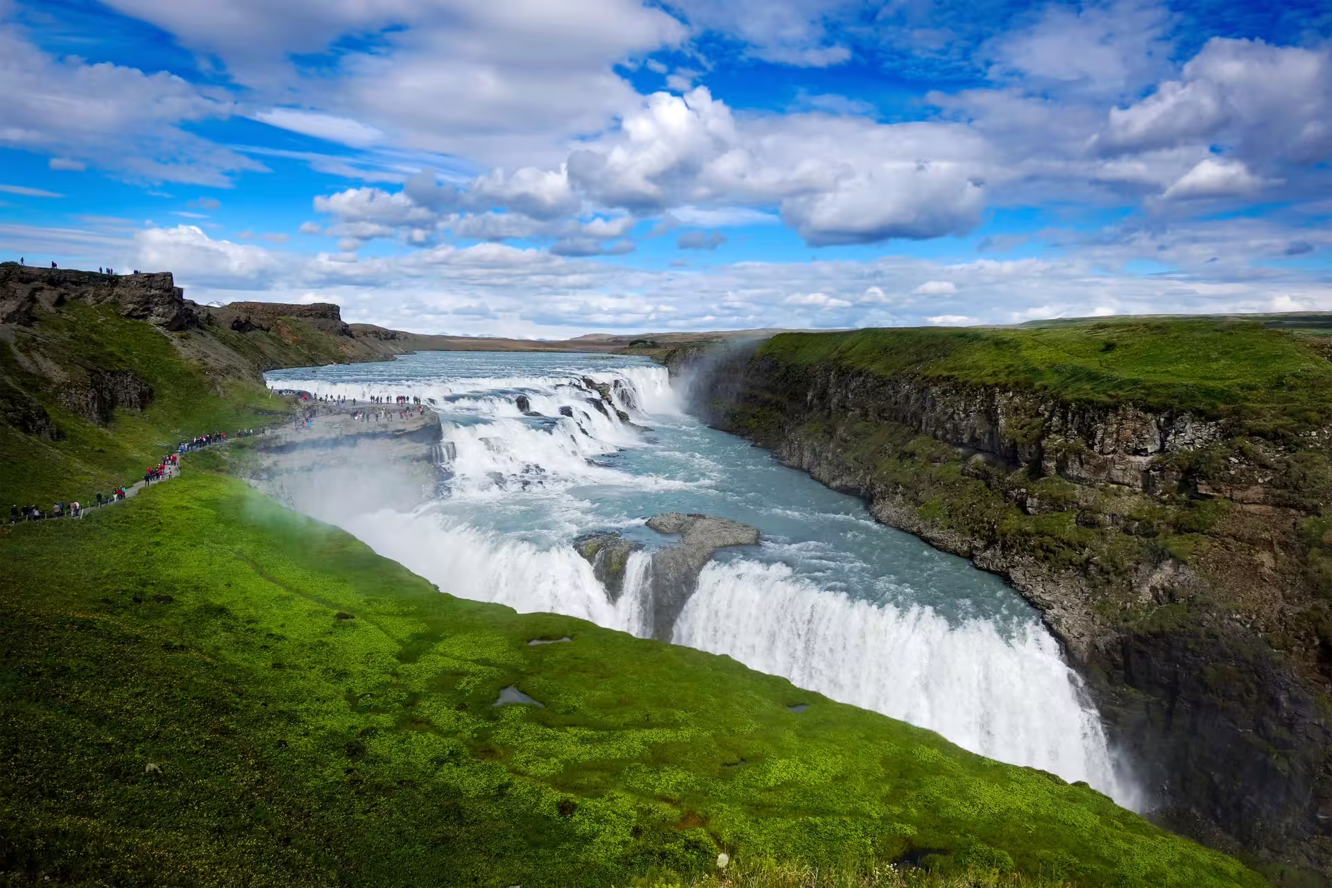 Breathtaking view of Gullfoss Waterfall cascading through lush green landscapes under a blue sky on the Golden Circle tour in Iceland.