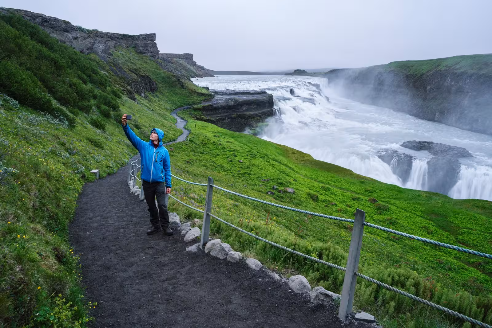 Tourist capturing a selfie by the majestic Gullfoss waterfall on a cloudy day in Iceland.