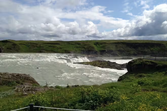 Churning white rapids of Gullfoss waterfall surrounded by green Icelandic landscape under cloudy sky