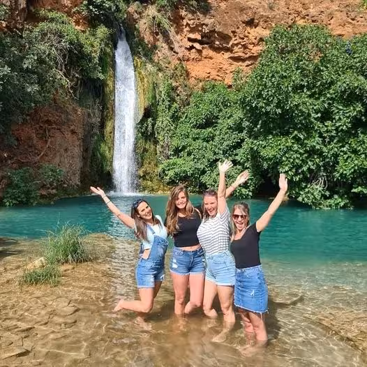 Four women posing in clear water beneath a hidden Algarve waterfall, a refreshing swimming stop on the half day jeep safari