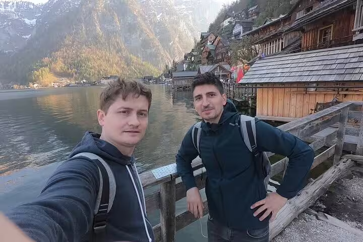 Two men taking a selfie on a wooden dock in Hallstatt with alpine lake and village in background