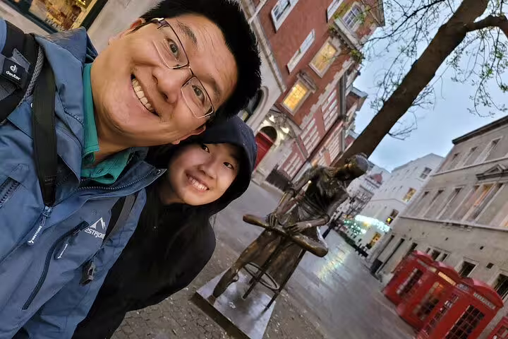 Father and child selfie with bronze statue on cobblestone street near red phone boxes in London