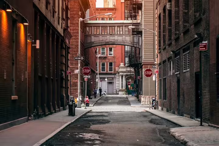 Narrow historic New York City street with brick buildings, stop signs, and a pedestrian bridge overhead