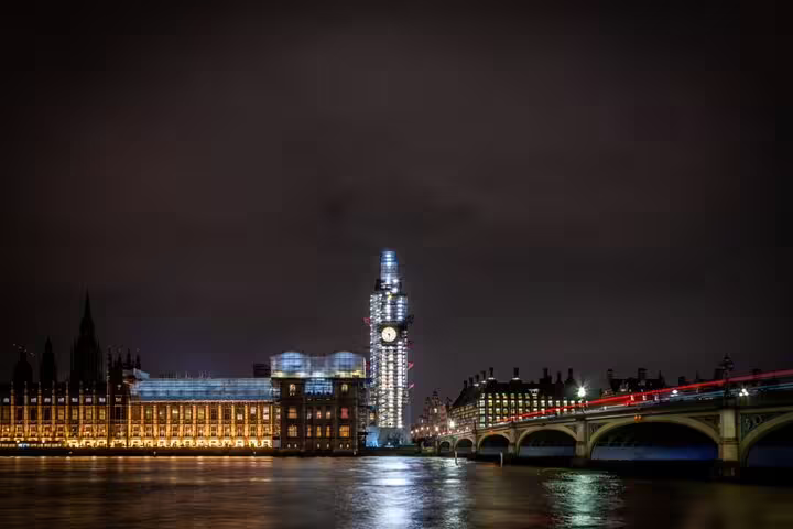 Big Ben clock tower under scaffolding at night with Westminster Bridge and River Thames, London