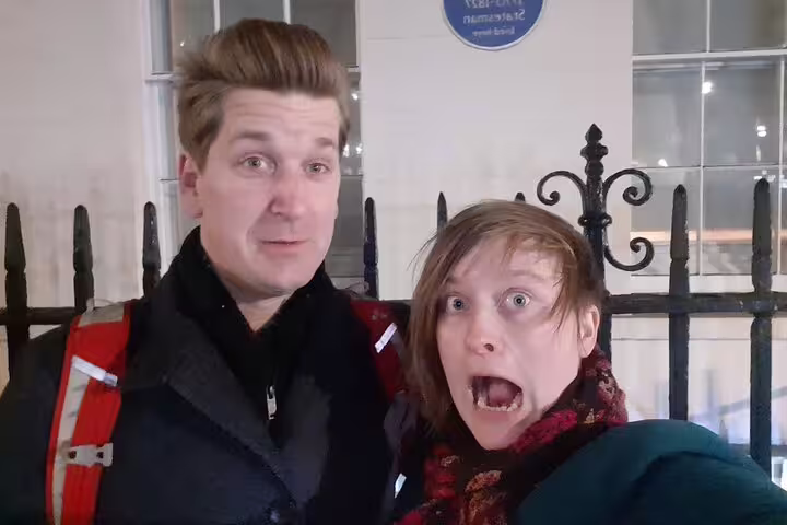 Two people making scared faces on a haunted London walking tour at night near iron gates