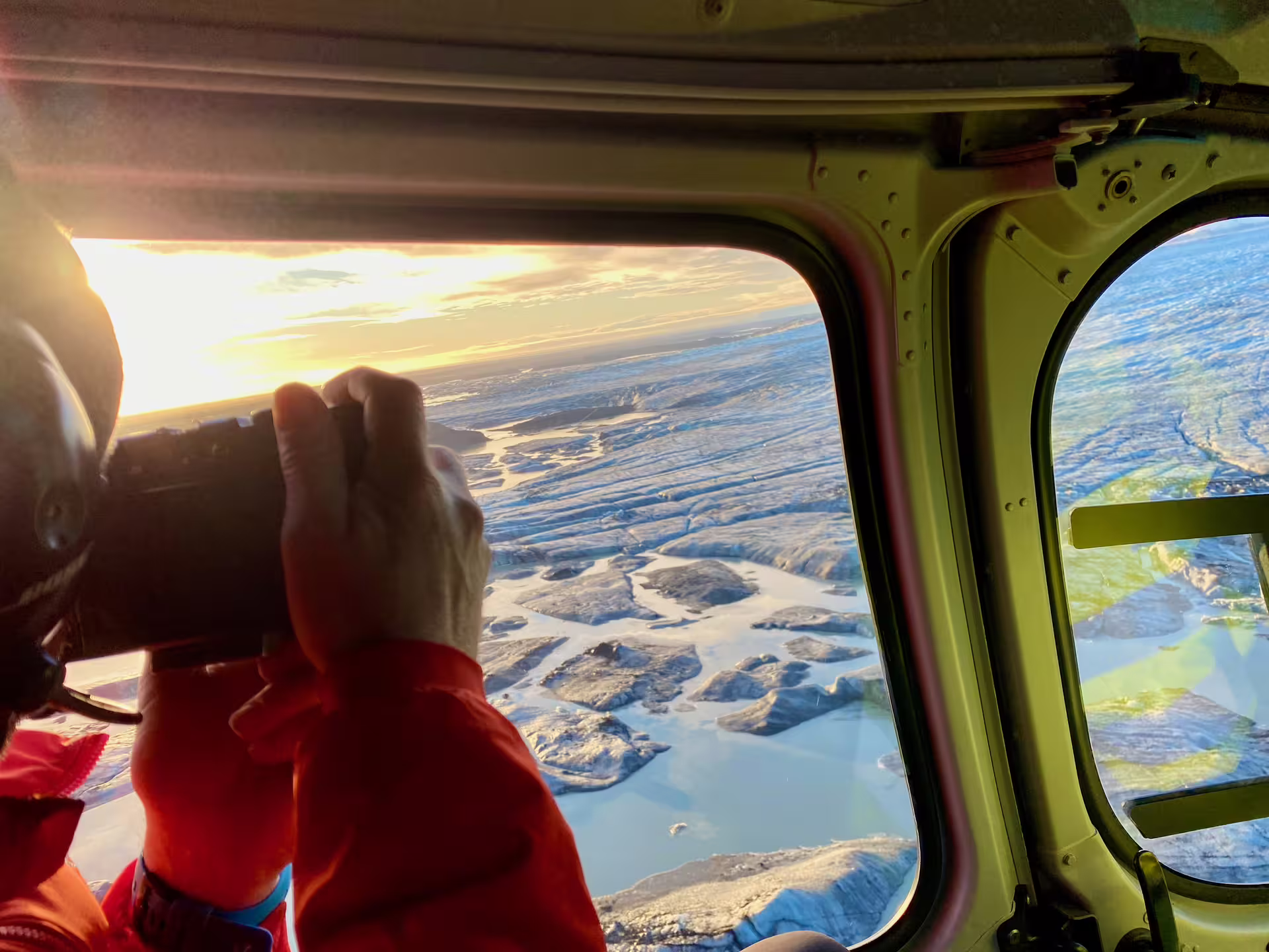 Photographer capturing aerial view of glacier and meltwater from helicopter window at golden sunset
