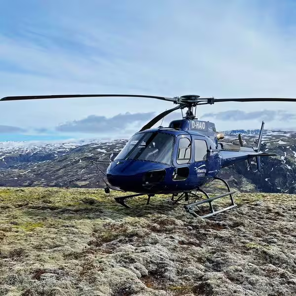 Blue helicopter D-HAIO landed on mossy highland terrain with snow-capped mountains in Iceland background