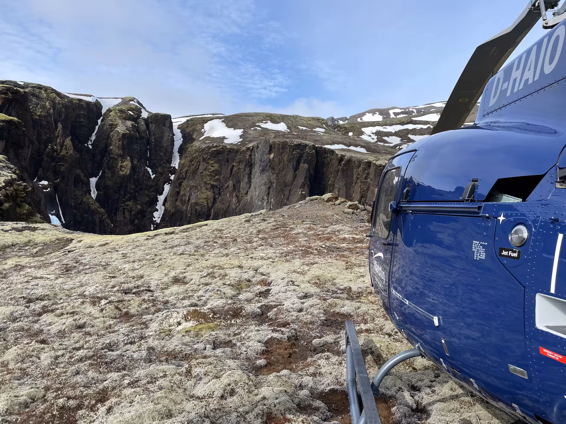 Blue helicopter landed on mossy tundra plateau near dramatic Iceland canyon cliffs with snow patches