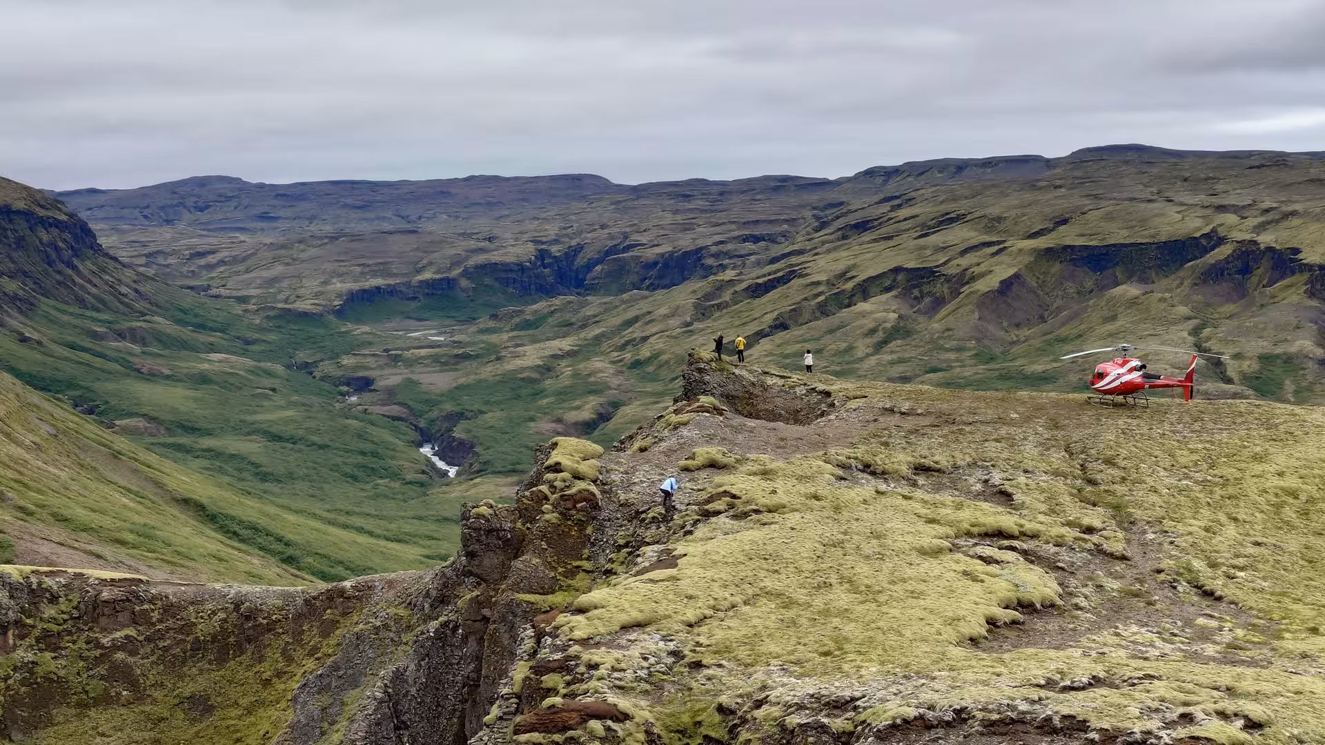 Red helicopter landed on mossy Iceland highland ridge with tourists overlooking a green canyon valley