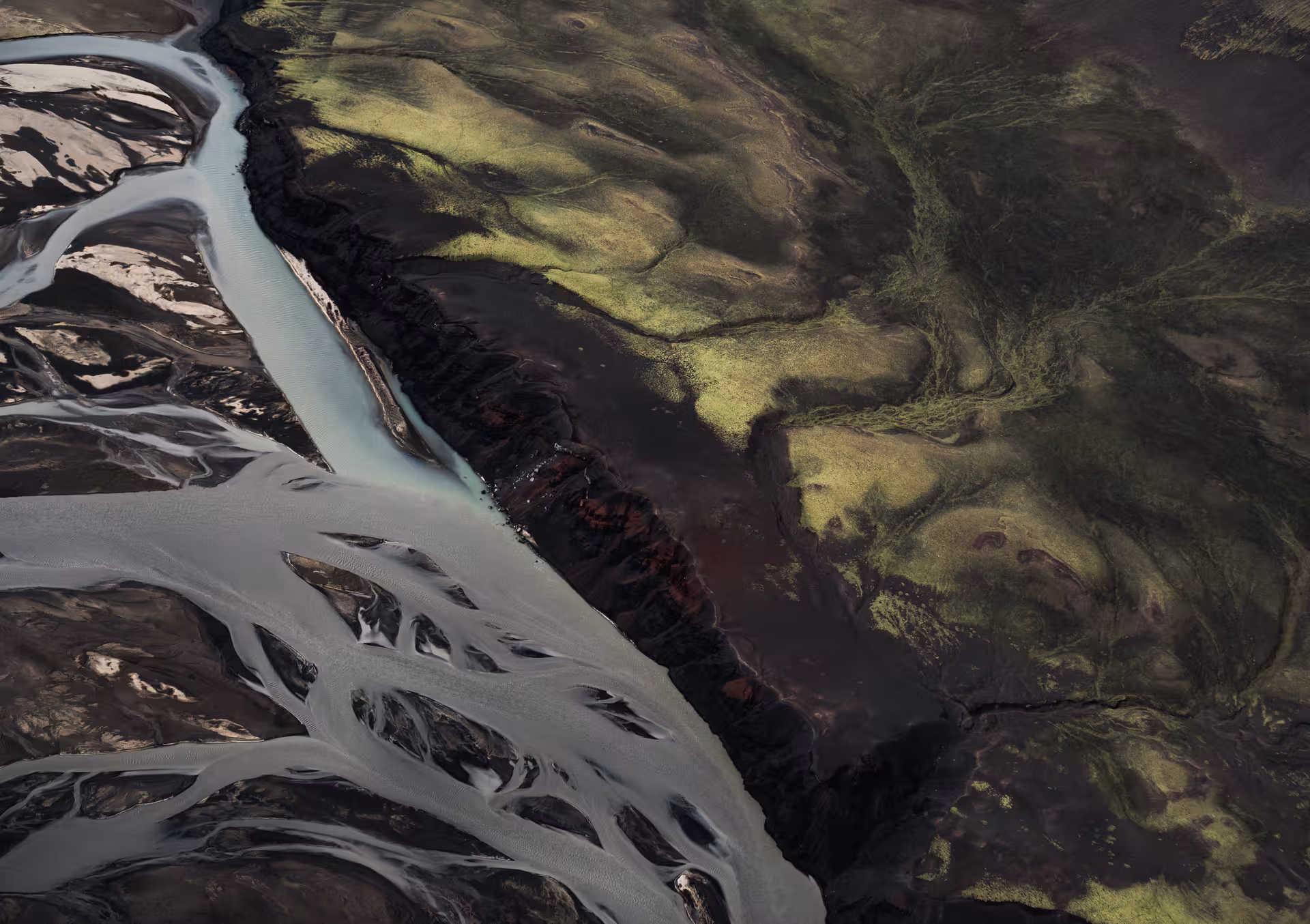 Aerial view of glacial braided rivers meeting green moss-covered volcanic terrain over Iceland's black sand plains