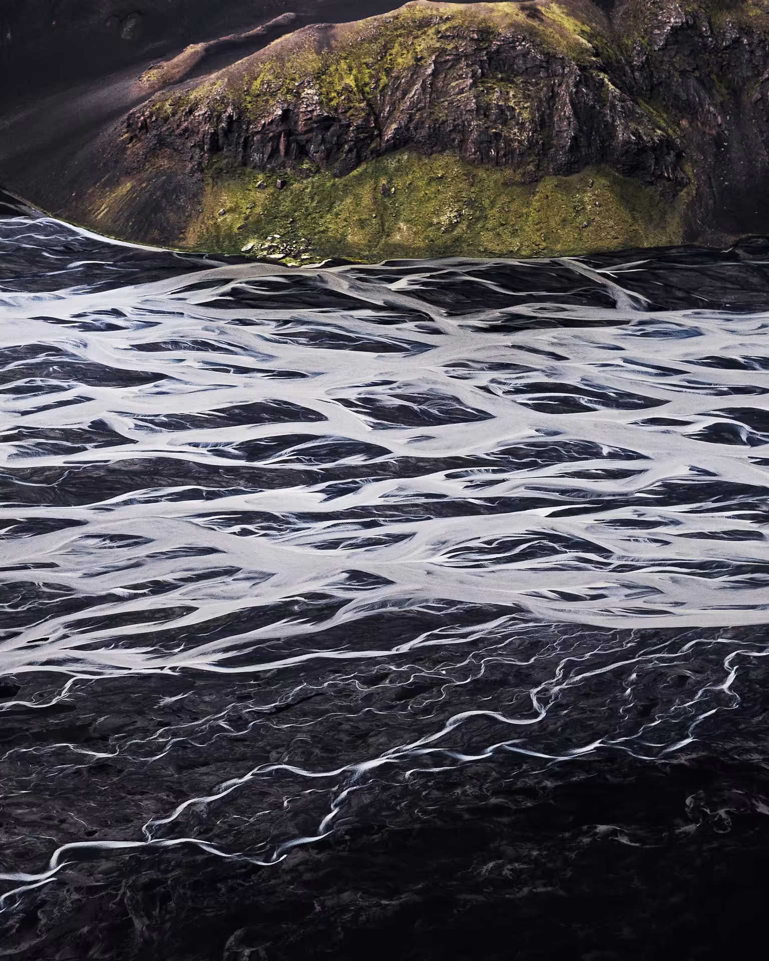 Aerial view of braided glacial rivers on black sand plains with moss-covered mountain in Iceland