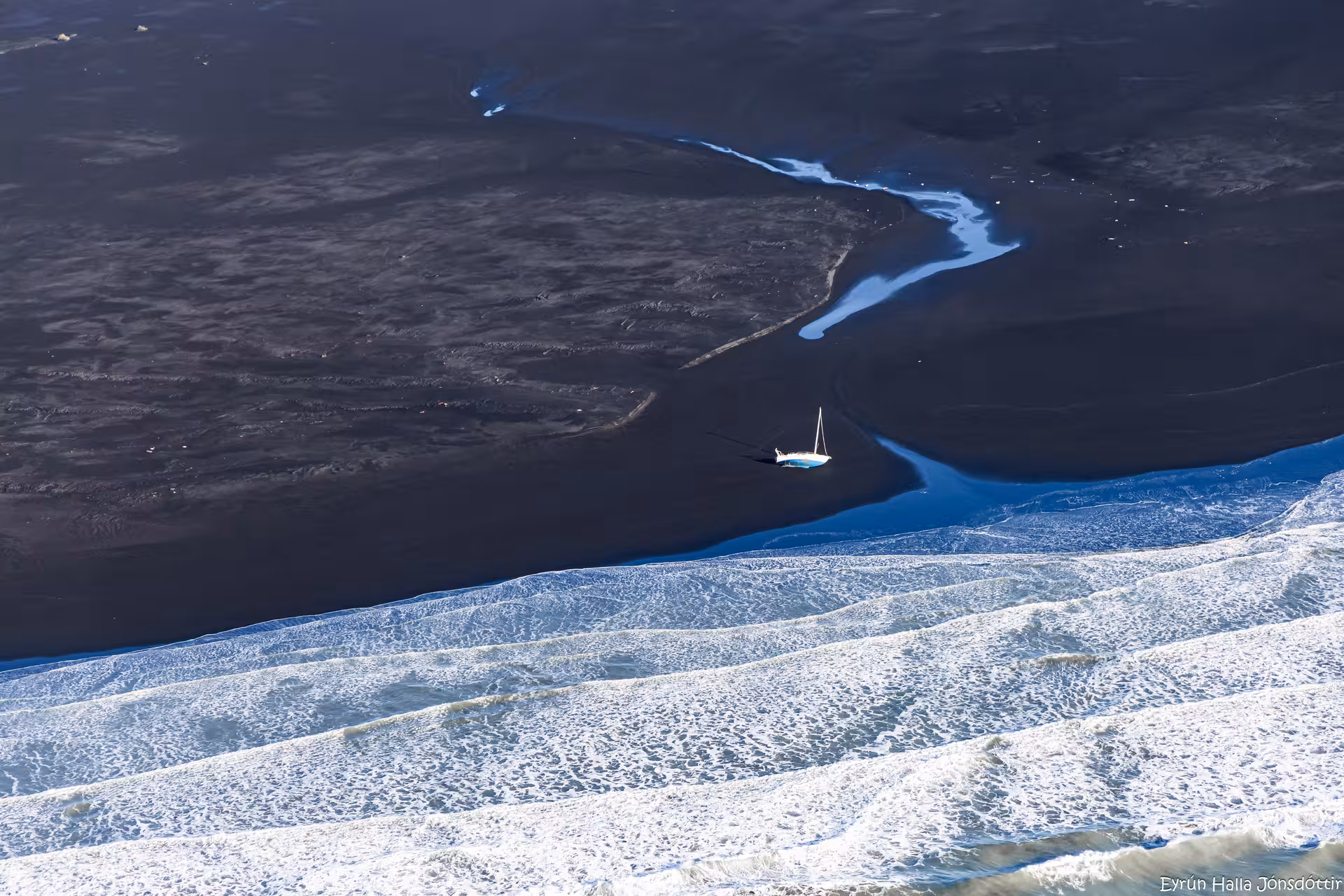 Aerial view of a sailboat stranded on Iceland's black sand plains with crashing ocean waves nearby